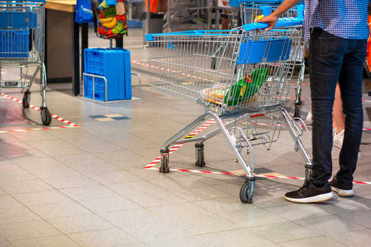 Man, With A Shopping Cart, Doing Groceries In A Supermarket During Coronavirus Covid-19 Pandemic. Tape On The Ground Indicates The Safe Social Distance. 