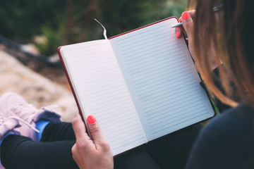 Side photo of blank notebook in female hands together with a pen, sitting in a park. Woman's hands...