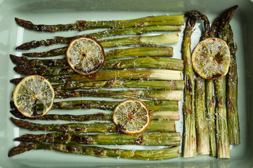Oven baked asparagus with lemon slices in ceramic dish, flat lay