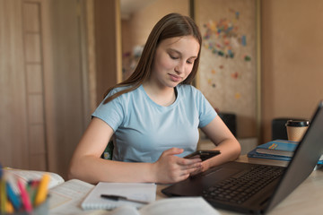 Teenager girl happy smiles, does homework on internet laptop, smartphone, social connections 4G 5G. Distance education training during quarantine. Background books lectures on school institute