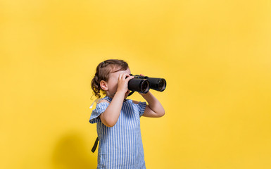 A little girl looks away with binoculars in her hands on a yellow background with space for text.