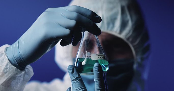 Close up of glass flask with liquid vaccine against coronavirus in hands of male Caucasian epidemiologist in full costume, mask, goggles and gloves. Laboratory testing of vaccines. Clinic research.