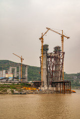 Fengdu, Chongqing, China - May 8, 2010: Yangtze River. Concrete suspension tower for bridge under construction with tall yellow cranes over brown water, cloudscape and green hill.