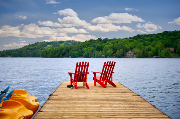 Two Muskoka chairs sitting on a wood dock facing a calm lake. Across the water is a cottage nestled...