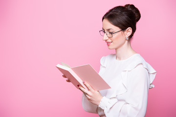 A young smiling woman in glasses with a notebook in her hands reads notes. Isolated on pink background