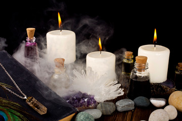 Magic potions, crystals, book, stones, peacock feather, and candles with smoke on a black background.