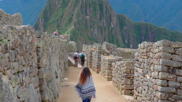 Slow motion tilt up of young woman wearing poncho walking amidst old ruins against mountains - Machu Picchu, Peru