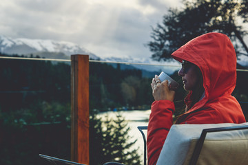 Young woman in vacation drinking coffee and thinking on a balcony in front of a beautiful lake with forest and mountain background.