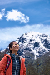Naklejka na ściany i meble Beautiful and colorful portrait in a cold winter of a smiling beautiful young woman wearing orange jacket and sunglasses. Surrounded by a dry forest with snow capped mountains behind and a blue sky