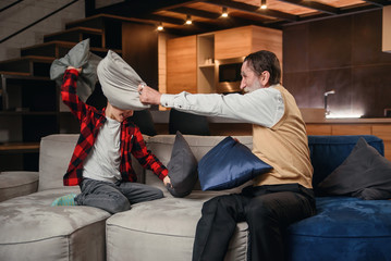 Happy boy with funny modern grandpa fighting with pillows having fun together sitting on...