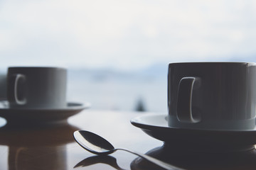 Taza de café sobre mesa de madera, con vista a lago, bosque y montañas.