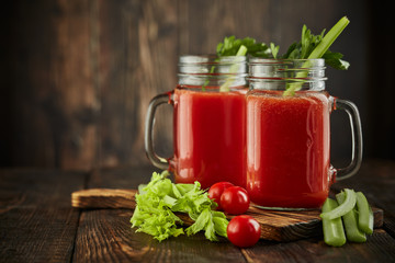 Two glasses with fresh tomato juice, celery, parsley and ripe tomatoes on dark brown wooden background.