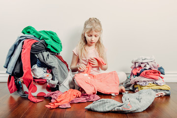 Mommy little helper. Cute Caucasian girl sorting clothes. Adorable funny child arranging organazing clothings. Kid with messy stack of clothes things on floor. Home chores housework for children.