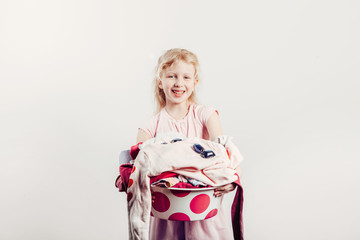 Mommy little helper. Smiling Caucasian girl child holding wash-basin with clothes and blue laundry detergent pods. Kid with messy stack of clothes. Home chores housework for children.