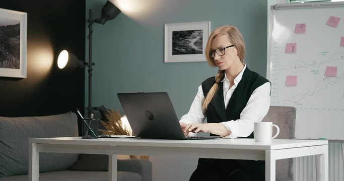 Competent businesswoman in formal outfit and glasses sitting at table and using wireless laptop. Mature lady with blond hair working on computer at office