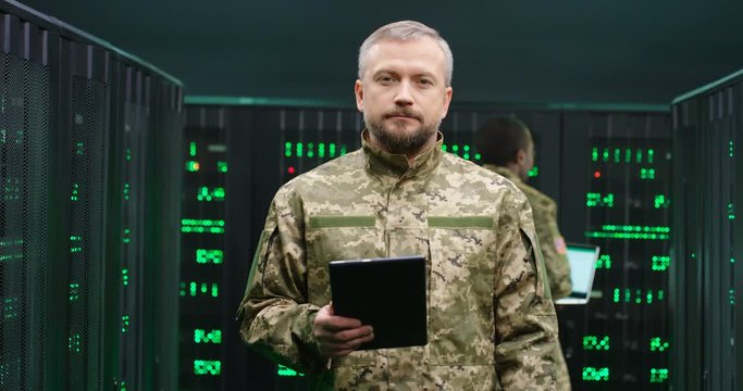 Portrait of serious Caucasian army official in camouflage uniform looking at camera and holding tablet device in dark server room. Analytic center concept. Military man posing in data center.