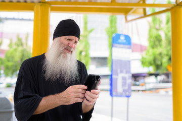 Mature handsome bearded hipster man using phone while sitting at the bus stop