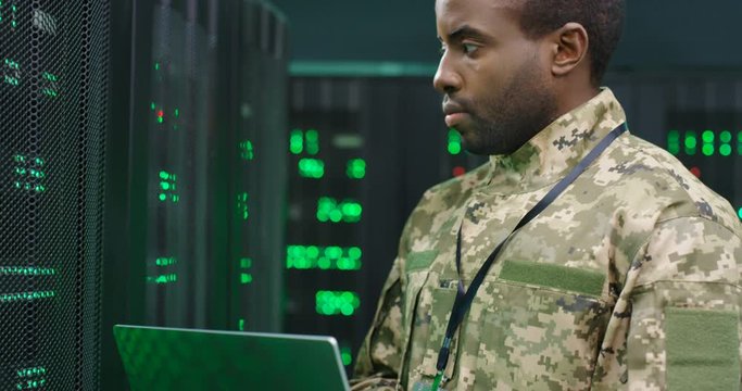 Close up of African American male army technician in camouglage costume and hat typing on keyboard of laptop computer while working with secret data at server room. Government analytic center.