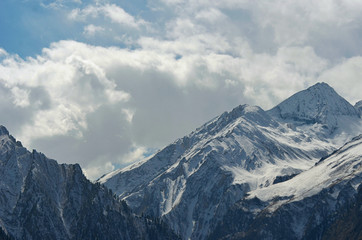 Steep mountain peaks are covered in snow. Some trees are on the lower slopes. Sunlight falls onto the snow, but the sky is filled with storm clouds.