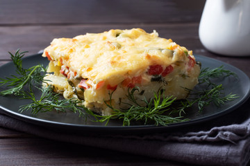Piece casserole on a plate of potatoes and vegetables with cheese. Dark wooden background. Close up.