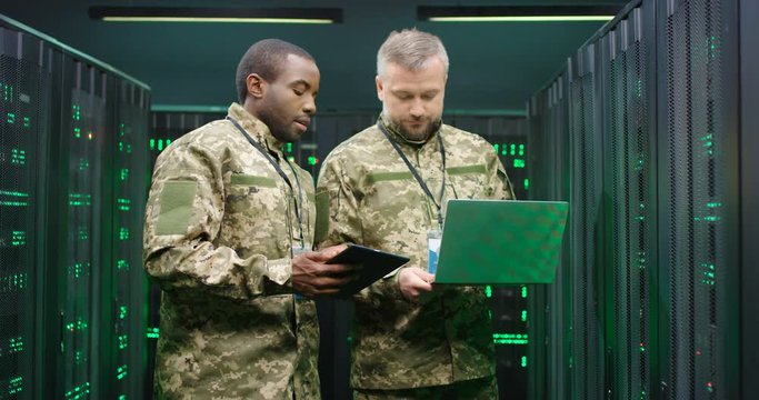 Young African American militarian with tablet device discussing some data and info with Caucasian senior officer with laptop computer. Monitoring server concept. Multiethnic male army colleagues.