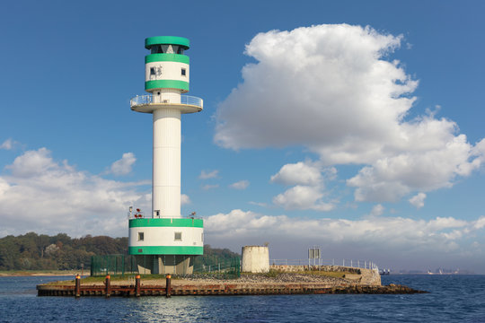 A sea-side close-up of the green and white lighthouse of Kiel-Friedrichsort in Germany. Sunshine, blue sky and clouds. In the background the entrance to the Baltic Sea.