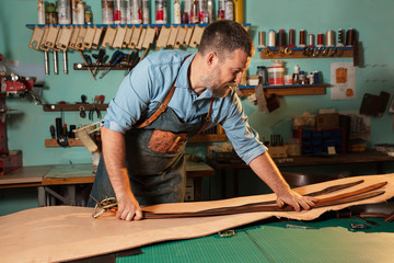 Craftsman in apron working with leather at workshop, belt manufacturing process.