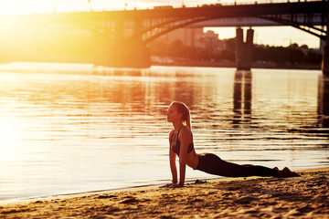 Woman practicing yoga meditation or stretching at sunrise on the sand. Morning yoga on the beach or coast of river on bridge urban city background