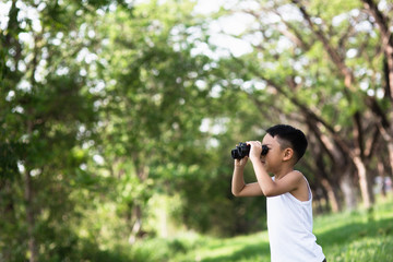 Little boy standing in a green grassy field scanning the surrounding woods with binoculars as he explores the countryside