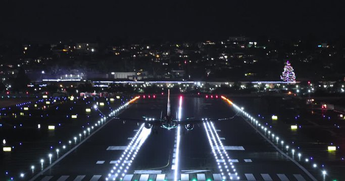Night footage of a plane landing on the runway at San Diego International Airport colorfully illuminated with red, green, white and blue lights. Night cityscape is seen on the background. 4K