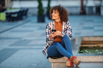 Young attractive mixed race woman sitting on fountain, using smart phone and enjoying music.