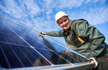 Cheerful male worker smiling to the camera while installing blue photovoltaic solar panel. Electrician in safety helmet under beautiful sky. Concept of alternative energy sources and innovations.