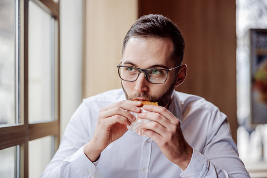 Young Man Sitting In Fast Food Restaurant Next To Window And Eating Cheese Burger.