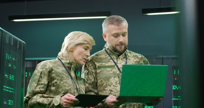 Caucsian female militarian with tablet device discussing some data and info with male officer with laptop computer. Monitoring server concept. Man and woman, army colleagues in digital center.