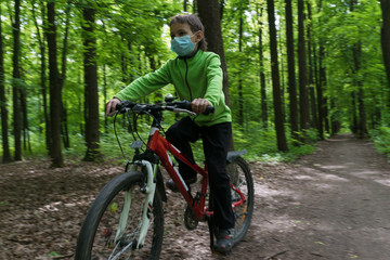 Fototapeta na wymiar boy in protective medical mask rides a bike in deep green summer forest, safe new way of sport activities after end of quarantine lockdown, outdoor sport activities