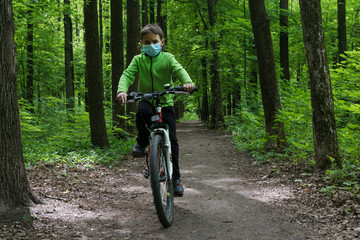 Naklejka premium boy in protective mask rides a bike in deep green forest, safe new way of sport activities after end of quarantine lockdown, outdoor sport activities