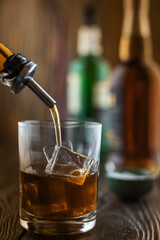 In the bar, whiskey is poured into a glass with ice, in the background there are bottles on a wooden table of the bar counter, shallow depth of field, selective focus. The concept of alcoholic drinks