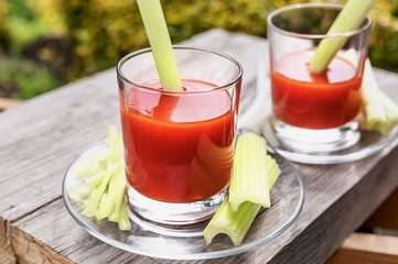 Two glasses of tomato juice with parsley and celery decorations, stand on wooden boards in the garden, morning sunlight shines, shallow depth of field, selective focus. The concept of natural drinks.