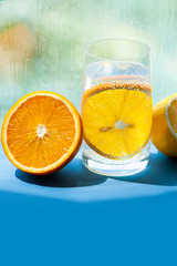 transparent glass with gaseous water and bubbles and a round slice of orange inside, half juicy and ripe orange and lemon next to it, glass with drops and smudges from the water on background