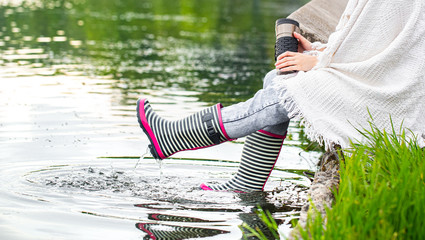 A young woman is sitting in rubber boots by the lake.
