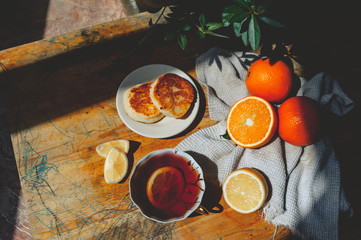 cozy tea party on a rustic wooden table, black tea with lemon, two cheesecakes on a saucer and ripe juicy oranges on a cotton napkin in the background in the harsh shadows of daylight