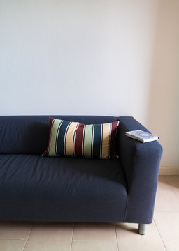 Sofa In Dark Blue With A Colorful Striped Throw Pillow And A Book On The Arm.  In A White Paint Wall Room.