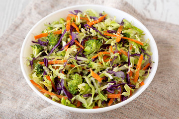 Homemade Raw Shredded Broccoli Slaw in a white bowl on a white wooden background, side view. Close-up.