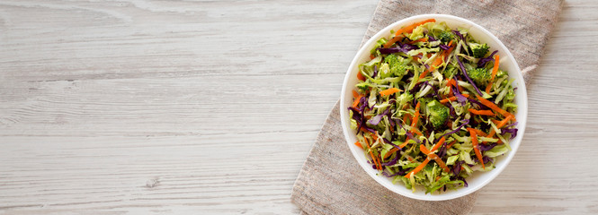 Homemade Raw Shredded Broccoli Slaw in a white bowl on a white wooden background, top view. Overhead, from above, flat lay. Space for text.