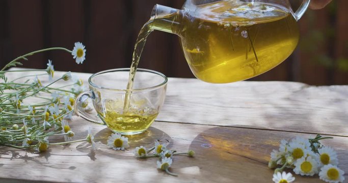 Chamomile tea is poured into a glass cup from a transparent teapot. The action takes place on a summer rural terrace with playing sunbeams. Nearby are the flowers of a pharmaceutical chamomile.