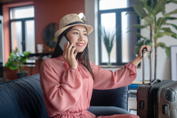 Dark-haired female in straw hat talking on mobile phone, sitting on sofa, holding suitcase handle