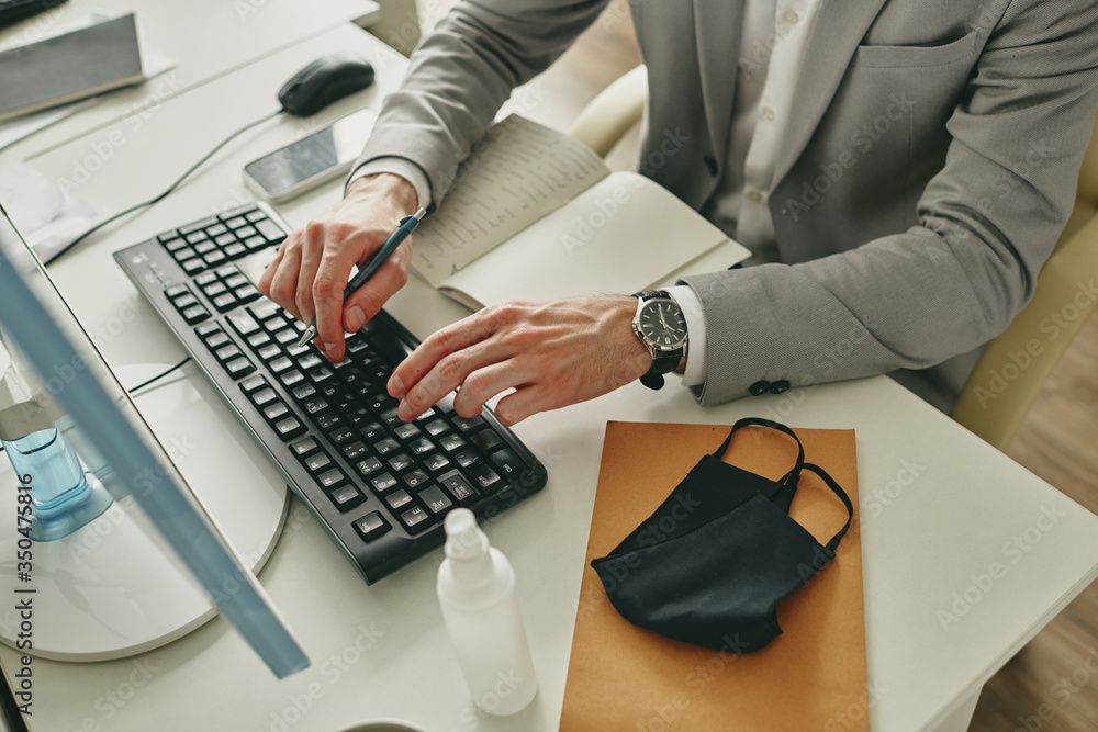 Aufkleber Close-up of unrecognizable businessman typing on computer keyboard while sitting at desk with facial mask and antiseptic bottle during quarantine