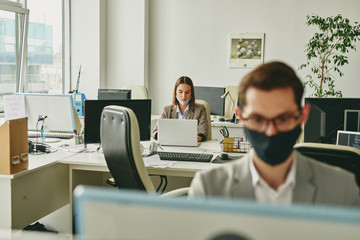 Young office woman with mask on chin using laptop while working in half-empty office during coronavirus pandemic