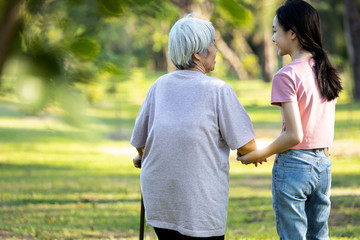 Back view of asian child girl hand holding her senior grandmother, walking together outside home,family time,old elderly taking a walk with her granddaughter in green nature background at the park