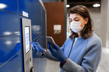 Fototapeta na wymiar health care, mail delivery and pandemic concept - woman wearing face protective medical mask for protection from virus disease with smartphone entering code on automated parcel machine's touch screen